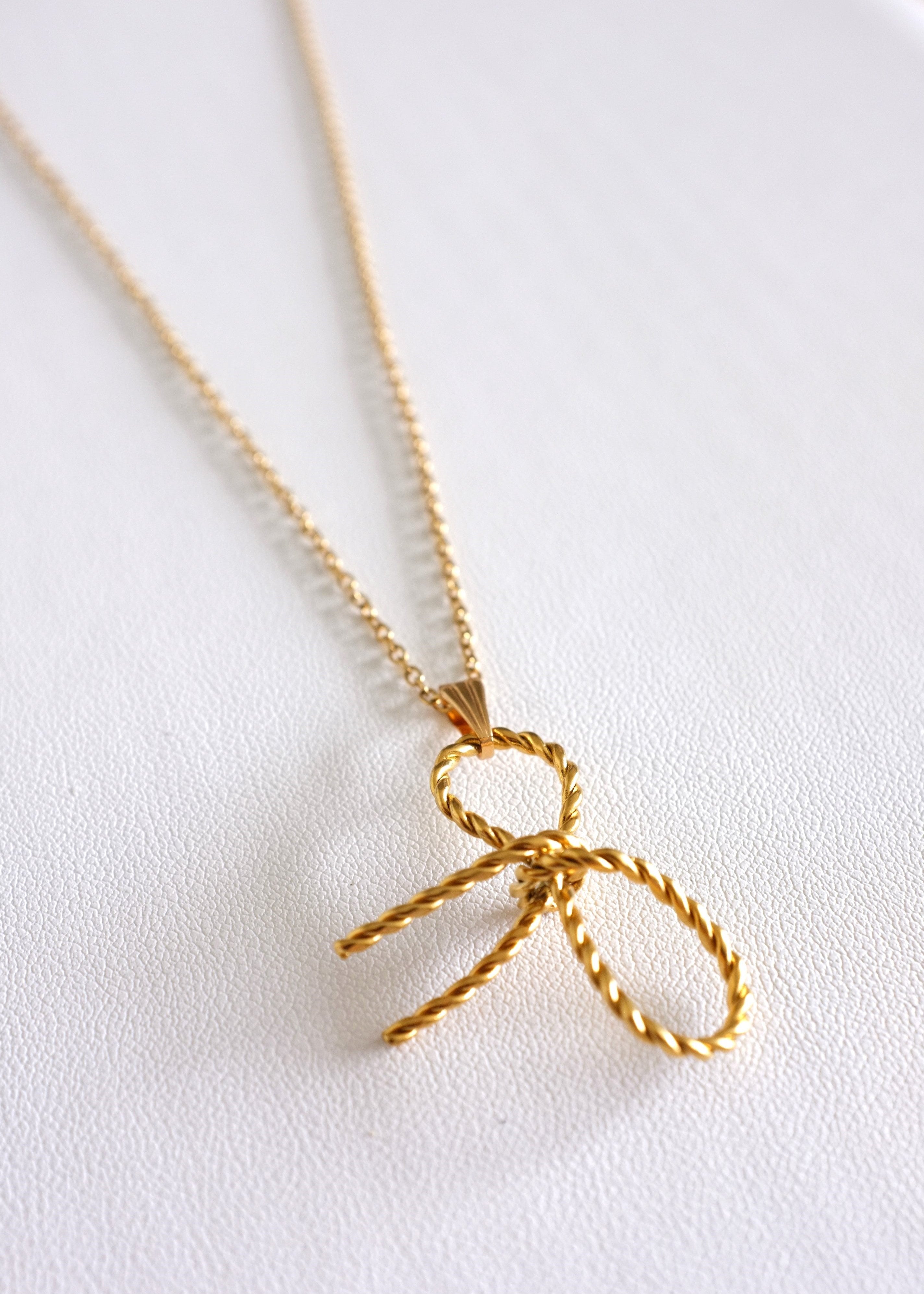 A gold-colored stainless steel necklace with a delicate bow pendant on a white background.