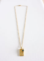 A close up of a stainless steel necklace with a unique rectangular pendant featuring a vibrant green gem, displayed against a white background.