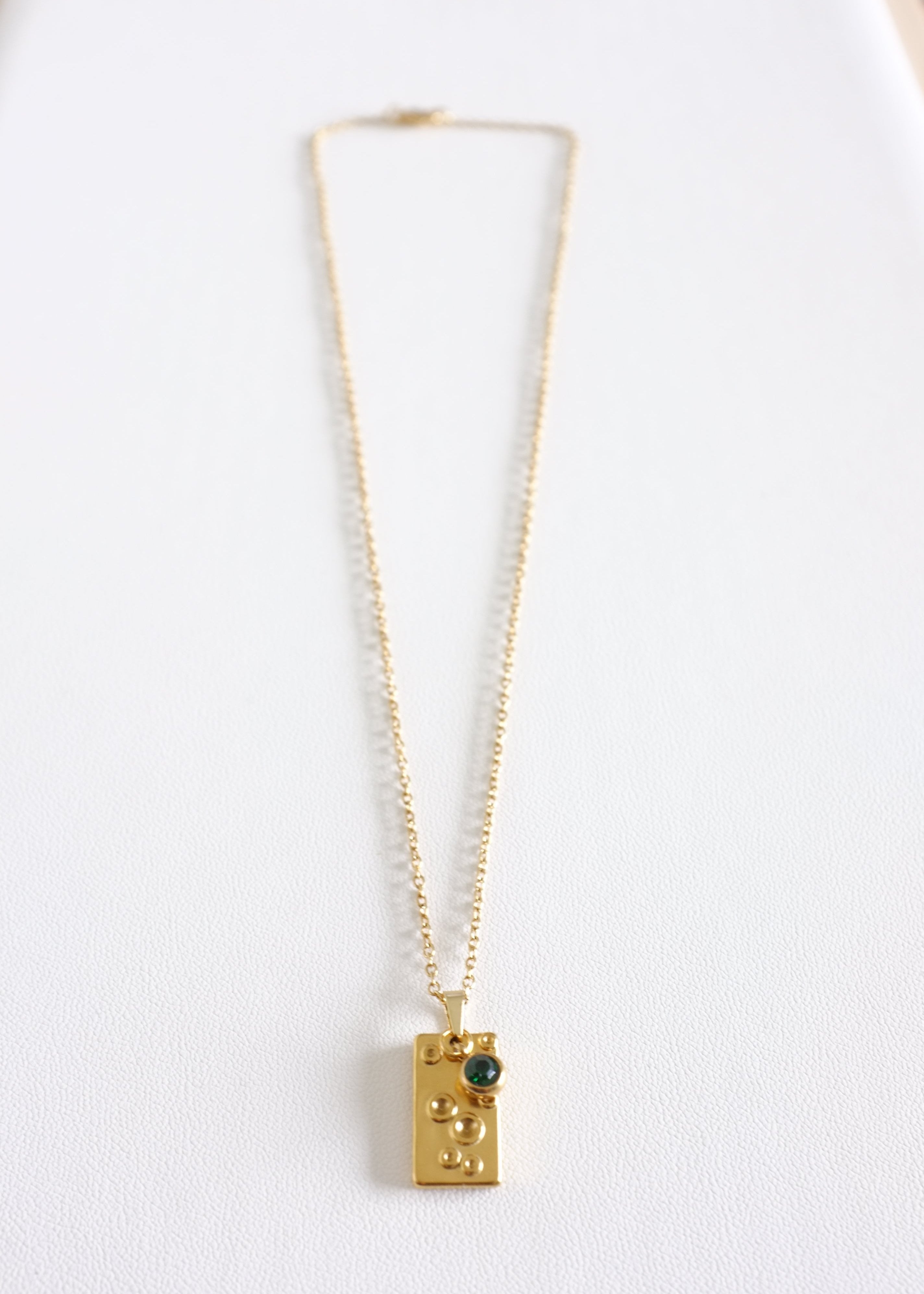 A close up of a stainless steel necklace with a unique rectangular pendant featuring a vibrant green gem, displayed against a white background.