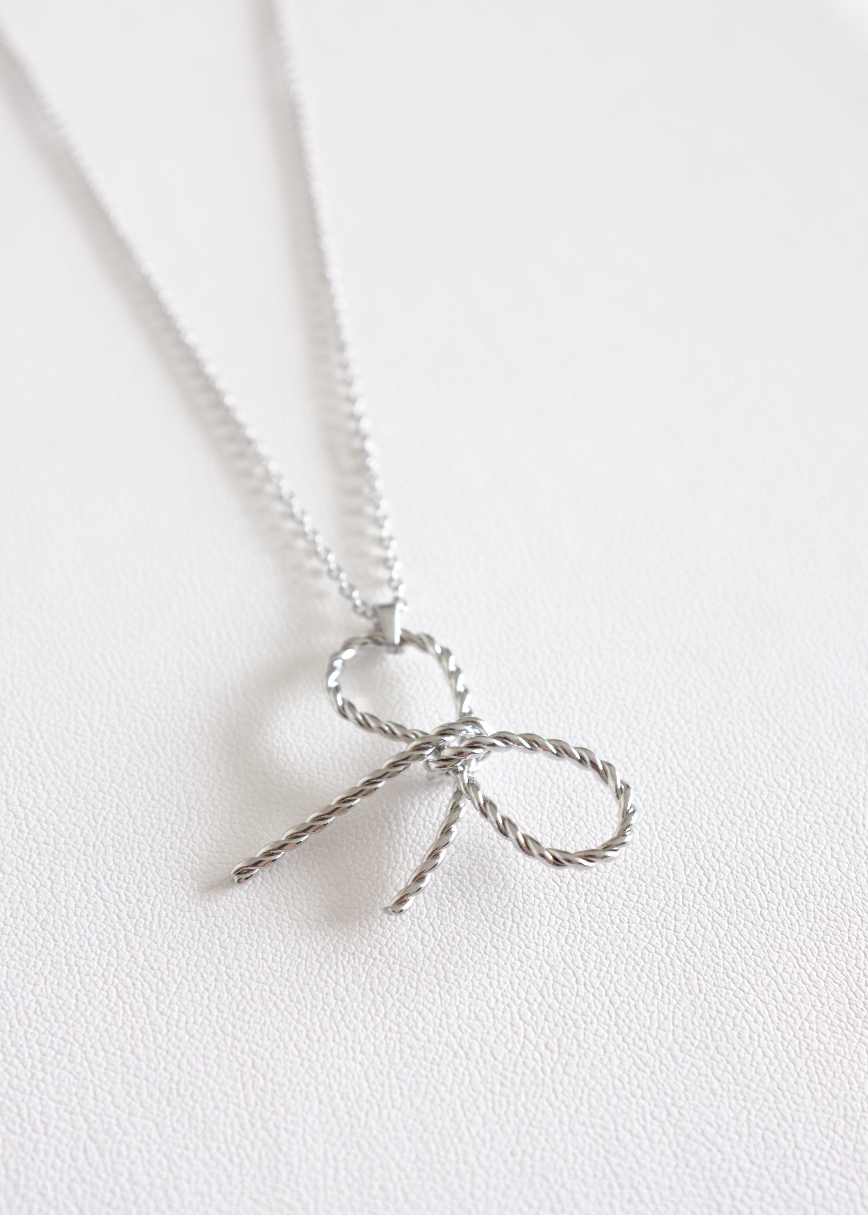 close up of a silver-colored stainless steel necklace with a delicate bow pendant on a white background.