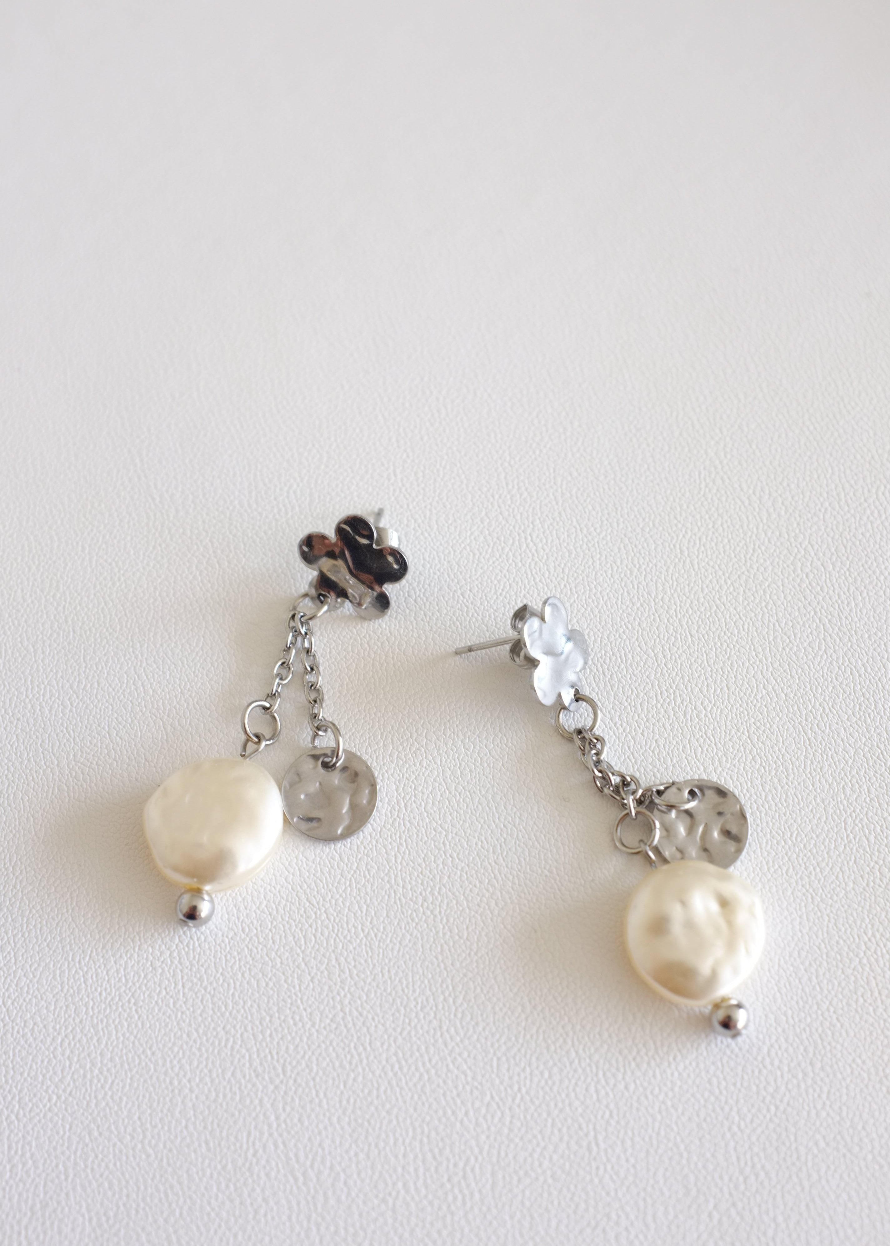 A pair of silver-colored floral stud earrings with a flat imitation pearl and a small pendant attached to two dainty chains on a white background