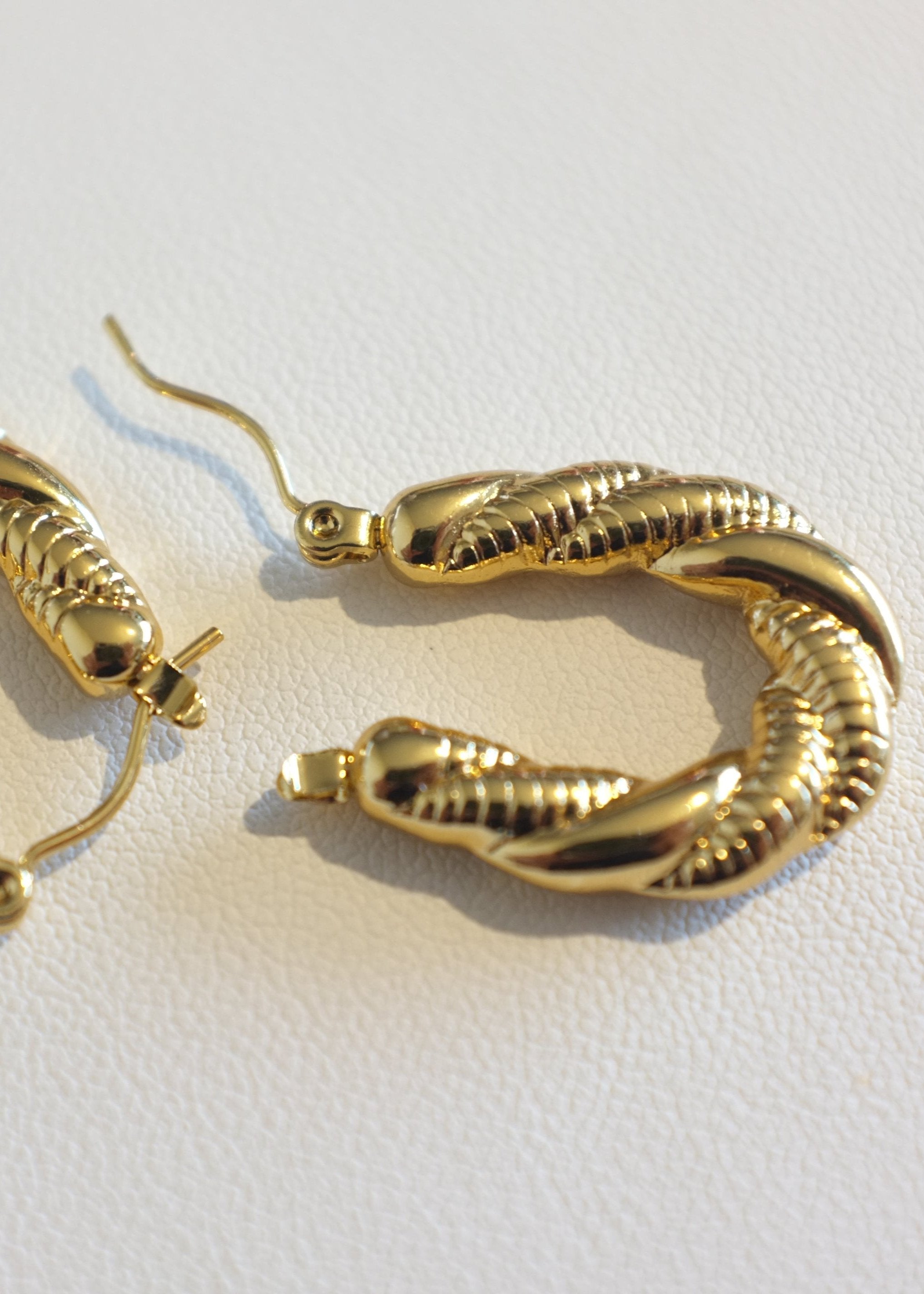 close up of a  pair of gold-colored stainless steel hoop earrings with a twisted rope design, displayed against a white background.