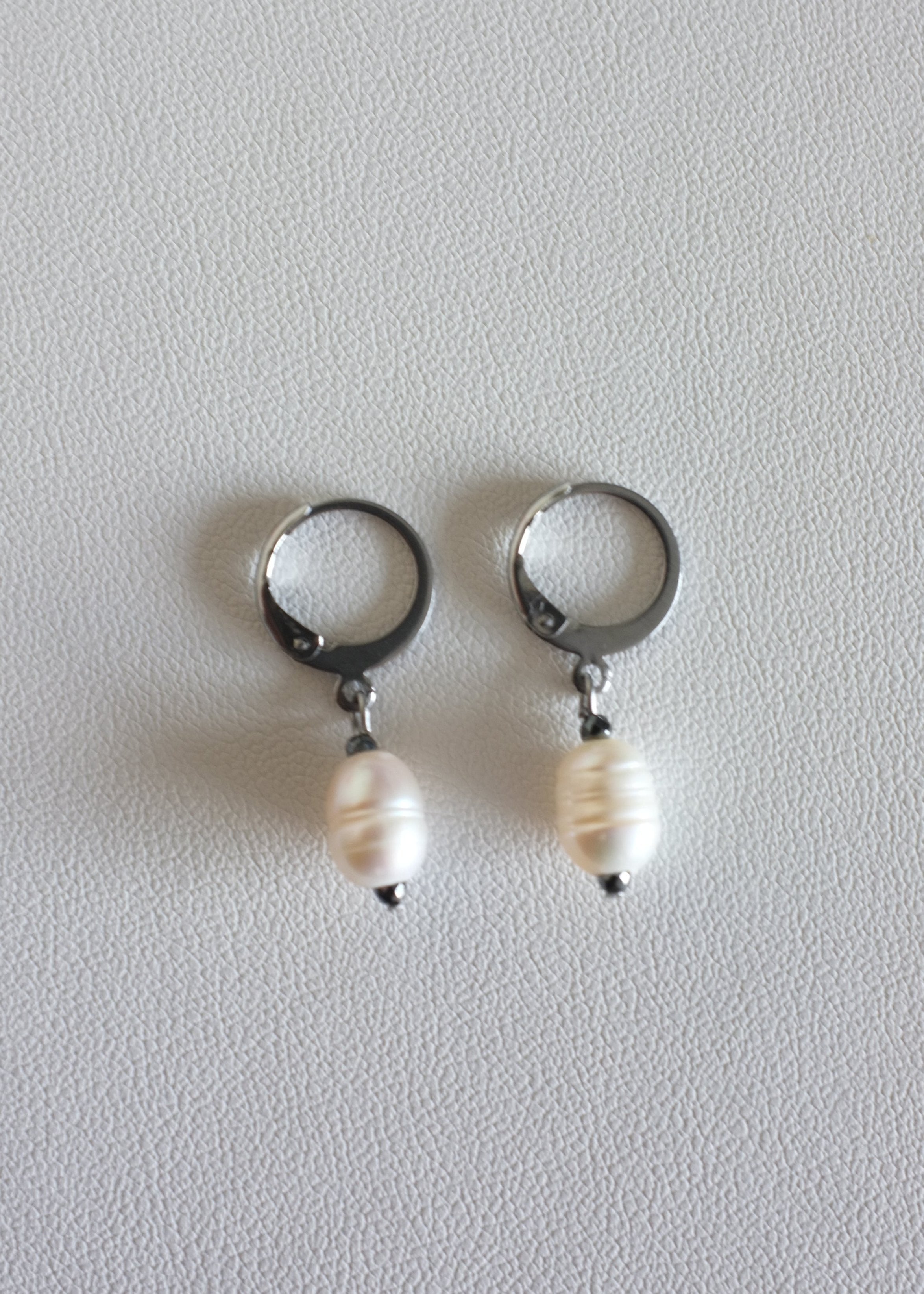 A pair of silver-colored stainless steel hoop earrings with freshwater pearl adornments on white background