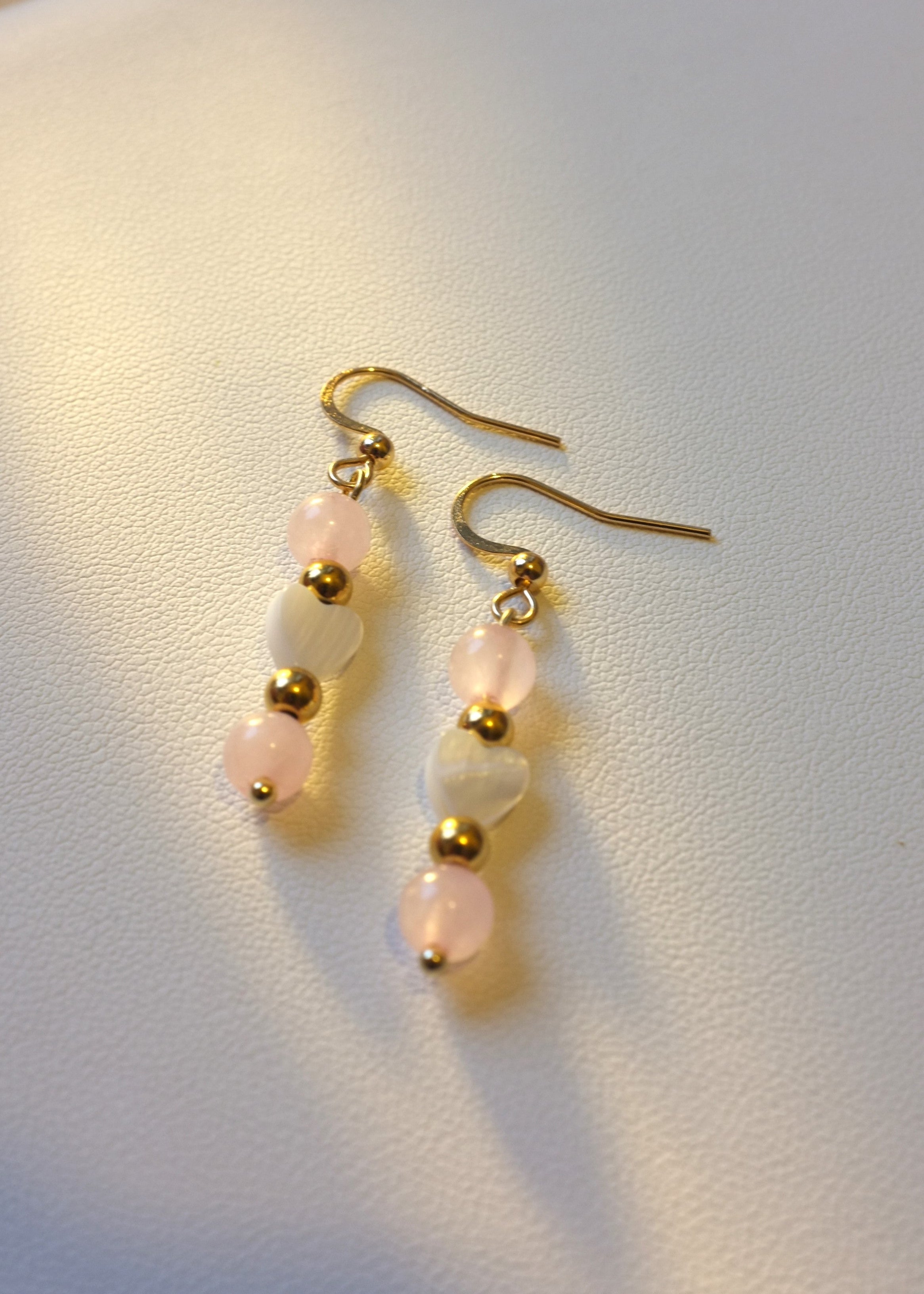 A pair of earrings featuring rose quartz stones and small heart-shaped shell accents on a gold-colored wire.