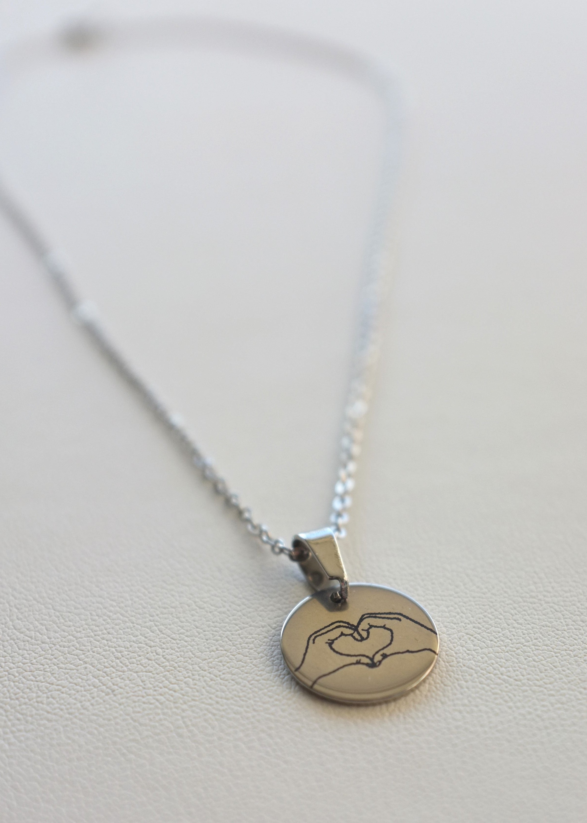 A close up of a stainless steel chain necklace with a round medal engraved with a heart symbol, displayed on a white background.