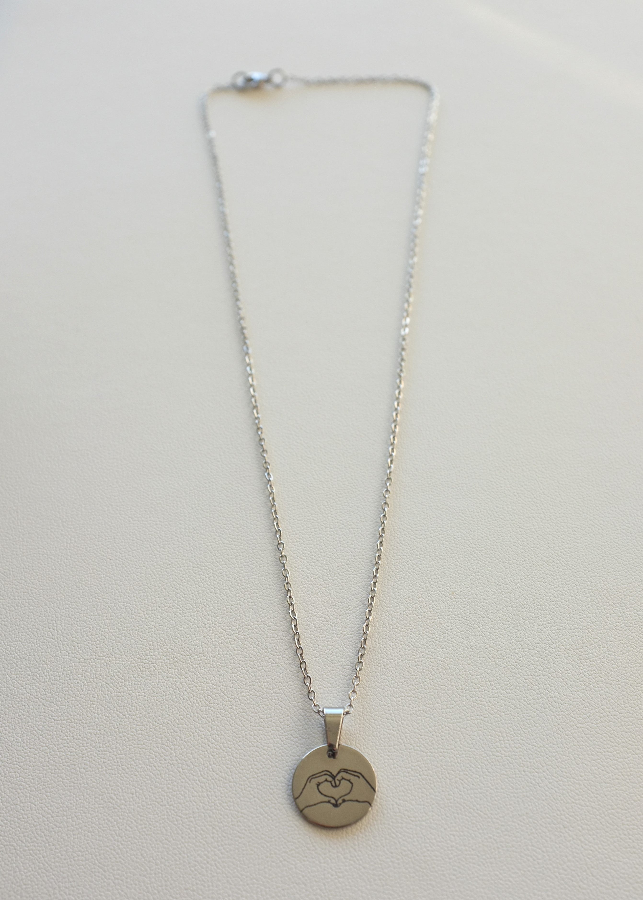 A stainless steel chain necklace with a round medal engraved with a heart symbol, displayed on a white background.